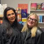 BBC Katie and Amy stand side by side and smile at the camera. Katie on the left has long, dark hair and is wearing a black jacket. On the right, Amy has shoulder-length, blonde hair and is wearing a pair of black framed glasses and a black leather jacket. Both are stood in front of a large bookshelf.