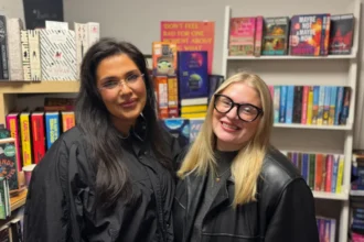 BBC Katie and Amy stand side by side and smile at the camera. Katie on the left has long, dark hair and is wearing a black jacket. On the right, Amy has shoulder-length, blonde hair and is wearing a pair of black framed glasses and a black leather jacket. Both are stood in front of a large bookshelf.