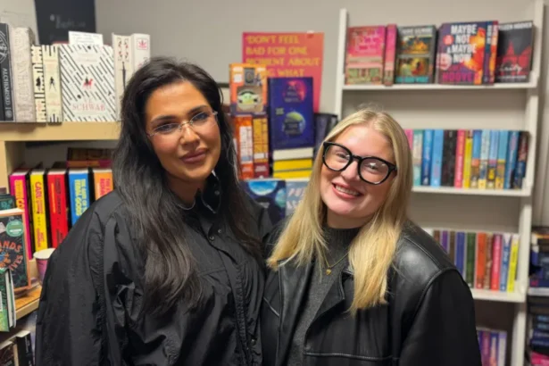 BBC Katie and Amy stand side by side and smile at the camera. Katie on the left has long, dark hair and is wearing a black jacket. On the right, Amy has shoulder-length, blonde hair and is wearing a pair of black framed glasses and a black leather jacket. Both are stood in front of a large bookshelf.