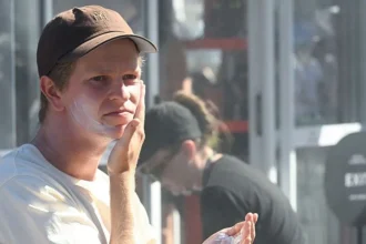 Getty Images A man wearing a white T-shirt and a brown baseball cap puts sunscreen on.