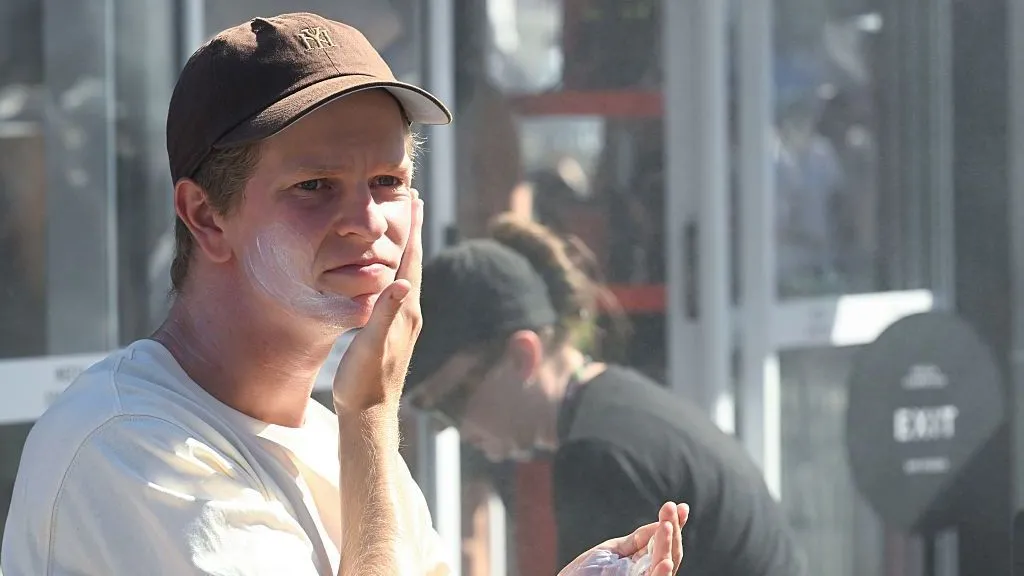 Getty Images A man wearing a white T-shirt and a brown baseball cap puts sunscreen on.