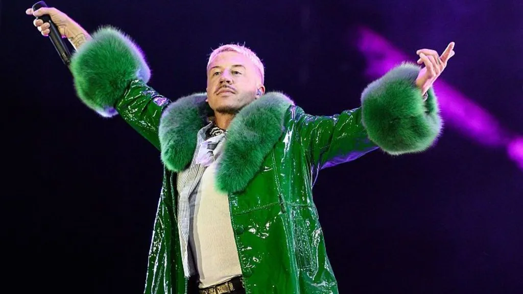05144a80-d12f-11f1-a94e-6dd2aca774ac-jpg Getty Images Macklemore, who has short, blond hair and short, dark stubble, raises his hands in the air as he holds a microphone while performing on stage. He wears a green coat with fur-type material around the neck and cuffs.