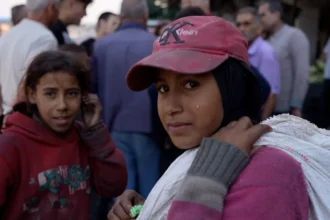 Neha Sharma/BBC A girl wearing a pink baseball cap with a white sack over her shoulder looks at the camera, with a second girl behind her and a crowd of people behind them standing further away