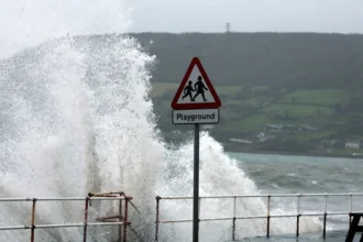 Reuters A wave crashing up onto a path with a metal railing and a black chair. There is a red and white triangular playground sign.