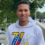 A man wearing a white hoodie and chain. The letter V in Venezuela's national colours and a map of the country is in the front of his hoodie. Behind him is a public street with cars and walkers in view.