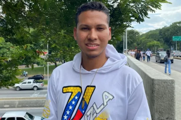 A man wearing a white hoodie and chain. The letter V in Venezuela's national colours and a map of the country is in the front of his hoodie. Behind him is a public street with cars and walkers in view.
