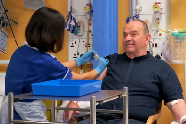 Addenbrooke's Hospital Tony Chapman sat in a hospital chair being given an injection. He is wearing a dark blue shirt. A a nurse in a blue uniform is giving him an injection of either the vaccine treatment or a placebo drug.