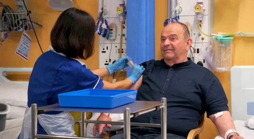 Addenbrooke's Hospital Tony Chapman sat in a hospital chair being given an injection. He is wearing a dark blue shirt. A a nurse in a blue uniform is giving him an injection of either the vaccine treatment or a placebo drug.