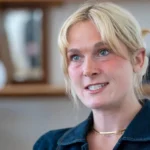 PA Media Lara Bird, who has blonde hair tied back, looking slightly off camera. She is wearing a denim blue top but only her head and shoulders are visible. She is standing in front of a blurred background of bookshelves.