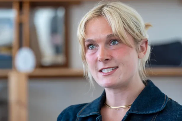 PA Media Lara Bird, who has blonde hair tied back, looking slightly off camera. She is wearing a denim blue top but only her head and shoulders are visible. She is standing in front of a blurred background of bookshelves.