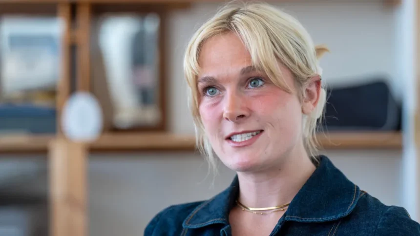 PA Media Lara Bird, who has blonde hair tied back, looking slightly off camera. She is wearing a denim blue top but only her head and shoulders are visible. She is standing in front of a blurred background of bookshelves.
