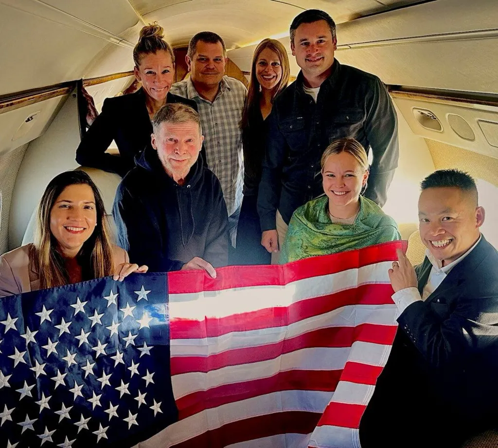 US Government / Handout via REUTERS Stephen Hubbard (second from left in the first row) poses with family members and American officials on board a flight to the US after his release from prison. Photo: 21 October 2026