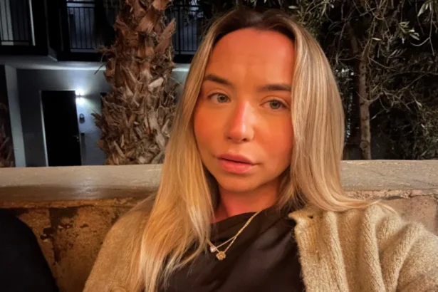 Hollie Sharman A woman with long blonde hair, she is wearing a beige cardigan, black top and a gold necklace. The background is trees and an apartment complex.