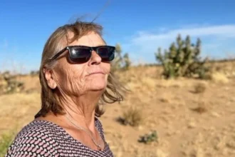 Christoph Greztmacher Eva Greztmacher looks up in sunglasses. A blue sky and desert scrubland can be seen behind her. She is wearing a necklace with a ring as a pendant.