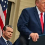 Getty Images Pete Hegseth seated while President Trump stands at the lectern in a blue suit and red tie