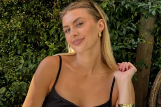 BBC Young woman with long blonde hair smiling, wearing a black dress, with green foliage in the background.