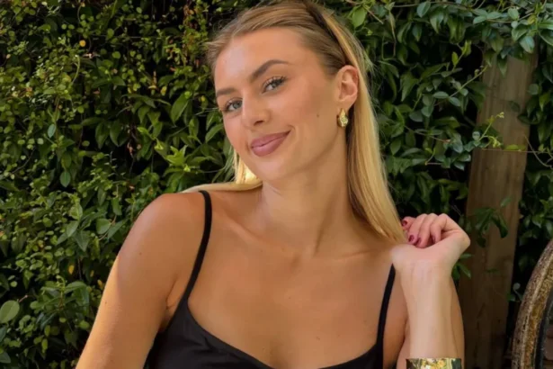 BBC Young woman with long blonde hair smiling, wearing a black dress, with green foliage in the background.