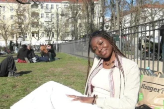 Amaka Osu A young woman with long braids is sitting on the grass in a park on a sunny day. She is tilted slightly to her left, smiling gently toward the camera. She is wearing  a cream top, jacket and trousers. Behind her is a paper bag with the word Hunger on it. In the background is a group of young people sitting on the grass