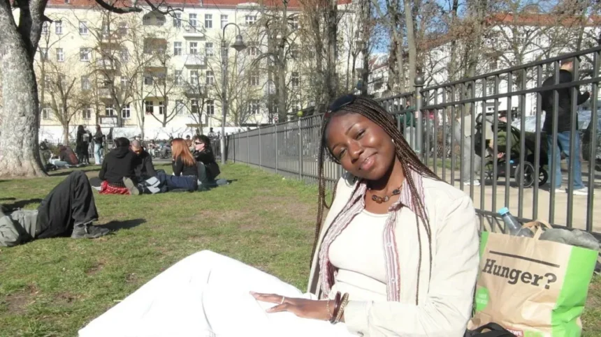 Amaka Osu A young woman with long braids is sitting on the grass in a park on a sunny day. She is tilted slightly to her left, smiling gently toward the camera. She is wearing  a cream top, jacket and trousers. Behind her is a paper bag with the word Hunger on it. In the background is a group of young people sitting on the grass
