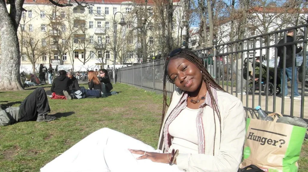 Amaka Osu A young woman with long braids is sitting on the grass in a park on a sunny day. She is tilted slightly to her left, smiling gently toward the camera. She is wearing  a cream top, jacket and trousers. Behind her is a paper bag with the word Hunger on it. In the background is a group of young people sitting on the grass