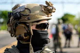 Reuters a US Immigration and Customs Enforcement officer standing outside of a detention center in New Jersey wearing a tactical helmet,  wrap-around sunglasses, and a cloth covering most of his face.