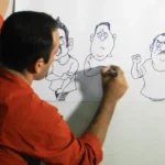Satish Acharya Satish Acharya wearing a rust shirt draws cartoons on a piece of paper attached to a white board