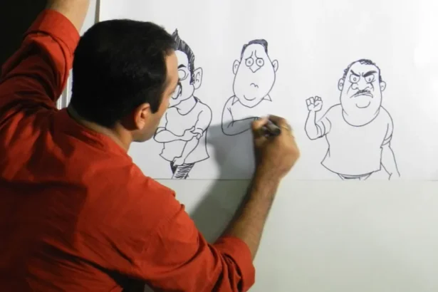 Satish Acharya Satish Acharya wearing a rust shirt draws cartoons on a piece of paper attached to a white board