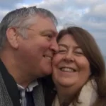 Louisa Nicoll A selfie of John kissing Louisa on the cheek they are both wrapped up warm in coats and scarves and smiling as the wind blows their hair with a blue cloudy sky and sea behind them