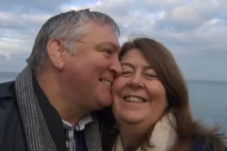 Louisa Nicoll A selfie of John kissing Louisa on the cheek they are both wrapped up warm in coats and scarves and smiling as the wind blows their hair with a blue cloudy sky and sea behind them