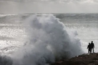 Getty Images A large wave crashes against a rock jutting out to sea on the coast of England. The sky is grey. There are two people standing on the rock.