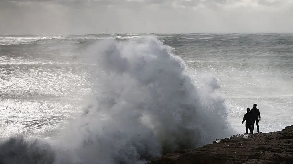 Getty Images A large wave crashes against a rock jutting out to sea on the coast of England. The sky is grey. There are two people standing on the rock.