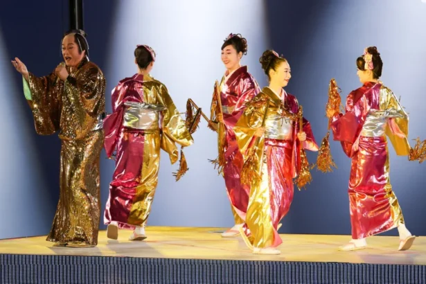 Getty Images A man and four women in shiny pink-gold traditional-style robes stand under bright stage lighting, holding decorative gold props - part of a performance at the Asian Games 2026 closing ceremony