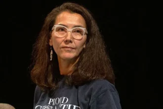 Getty Images Peltola looks to the side as she wears a grey T-shirt and white-framed glasses