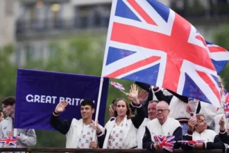 PA Media Tom Daley and other members of Team GB wave from a boat in Paris holding an enormous Union Jack flag aloft, with a Great Britain banner on purple cloth can be seen behind them