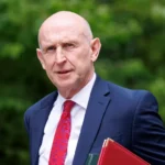 EPA/Shutterstock John Healey walks into Downing Street for a cabinet meeting. He is wearing a blue suit, red tie and white shirt and carries a red, and a green, folder under his arm.