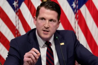 Getty Images A man in a navy-blue suit, white shirt, and blue-and-red striped tie speaks into a microphone. A small rectangular gold pin is attached to his lapel, with two American flags standing behind him.