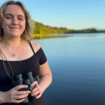 Nina Torrance/BBC Alba Caitlin Andrew is a young woman with shoulder length blonde hair. She is smiling as looks into the camera while holding a pair of black binoculars. Behind her is a loch with trees along its shores.
