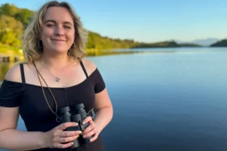 Nina Torrance/BBC Alba Caitlin Andrew is a young woman with shoulder length blonde hair. She is smiling as looks into the camera while holding a pair of black binoculars. Behind her is a loch with trees along its shores.