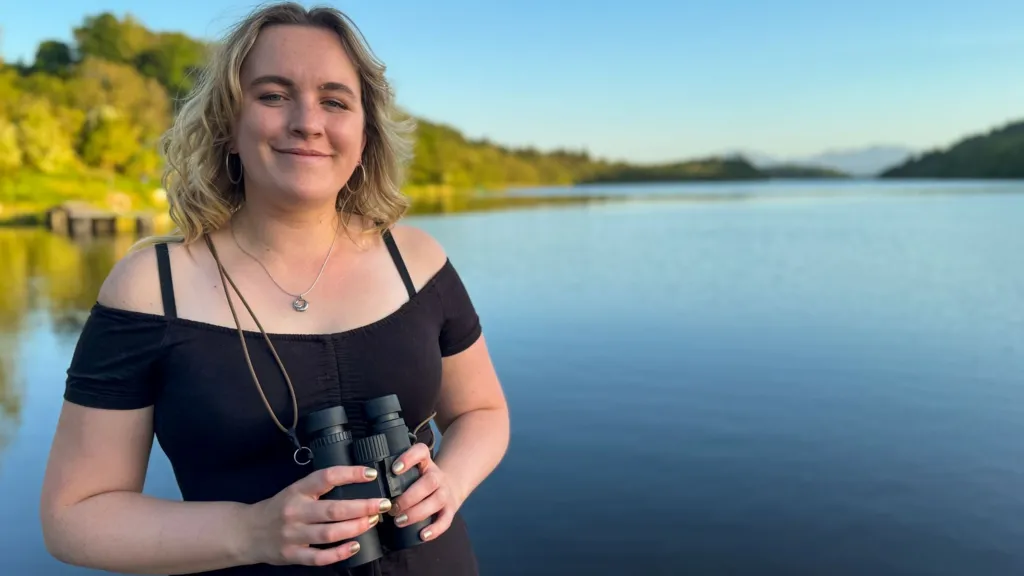 Nina Torrance/BBC Alba Caitlin Andrew is a young woman with shoulder length blonde hair. She is smiling as looks into the camera while holding a pair of black binoculars. Behind her is a loch with trees along its shores.