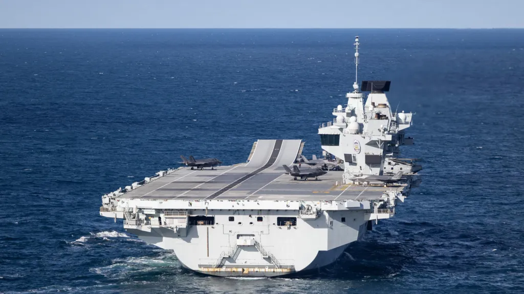 488b8820-cdb3-11f1-bc2e-018d645d8d21-jpg UK Ministry of Defence A large grey carrier is seen on the open ocean.