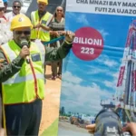 Tanzania's energy ministry Energy Minister Juma Homera in yellow hard hat and yellow vest holds a microphone by a big poster as he announces to a small group and a photographer the discovery of 223 billion feet of natural gas.