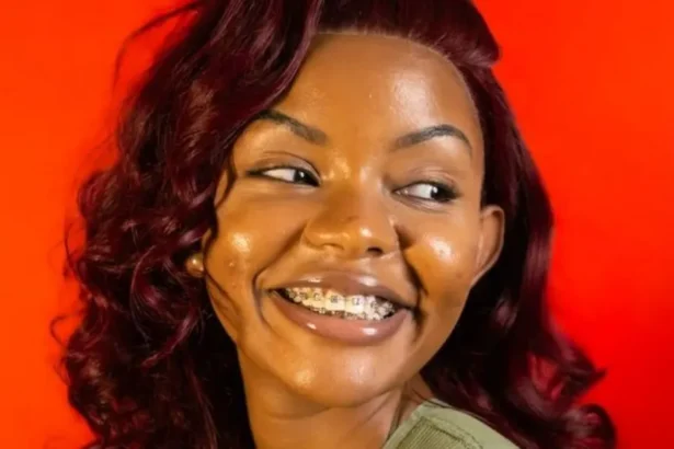 That Girl Boina A young woman in braces and a burgundy wig, wearing a green vest, smiling away from the camera with her head tilted to the left behind a red background