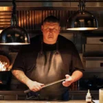 Apple TV Gareth Ward in black chef overalls holding large knife with kitchen appliances and open fire behind him and large overhead light pendants. He has a tattoo down one arm and wears a leather apron and has short dark hair.