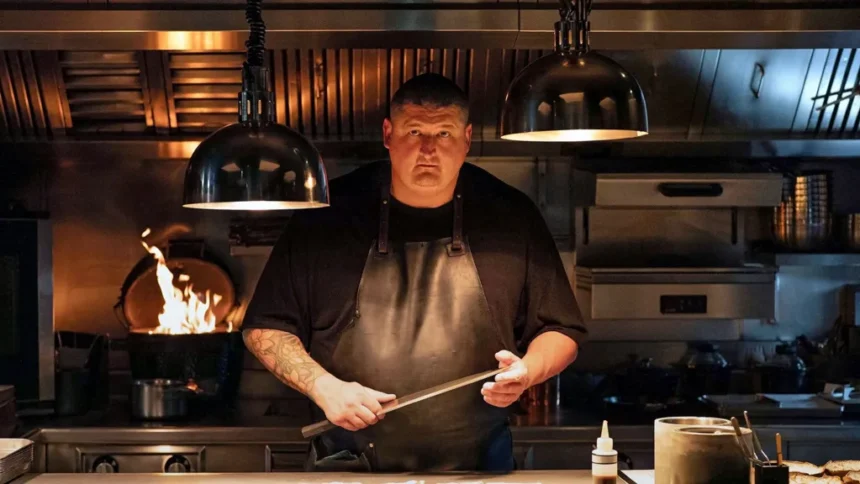 Apple TV Gareth Ward in black chef overalls holding large knife with kitchen appliances and open fire behind him and large overhead light pendants. He has a tattoo down one arm and wears a leather apron and has short dark hair.