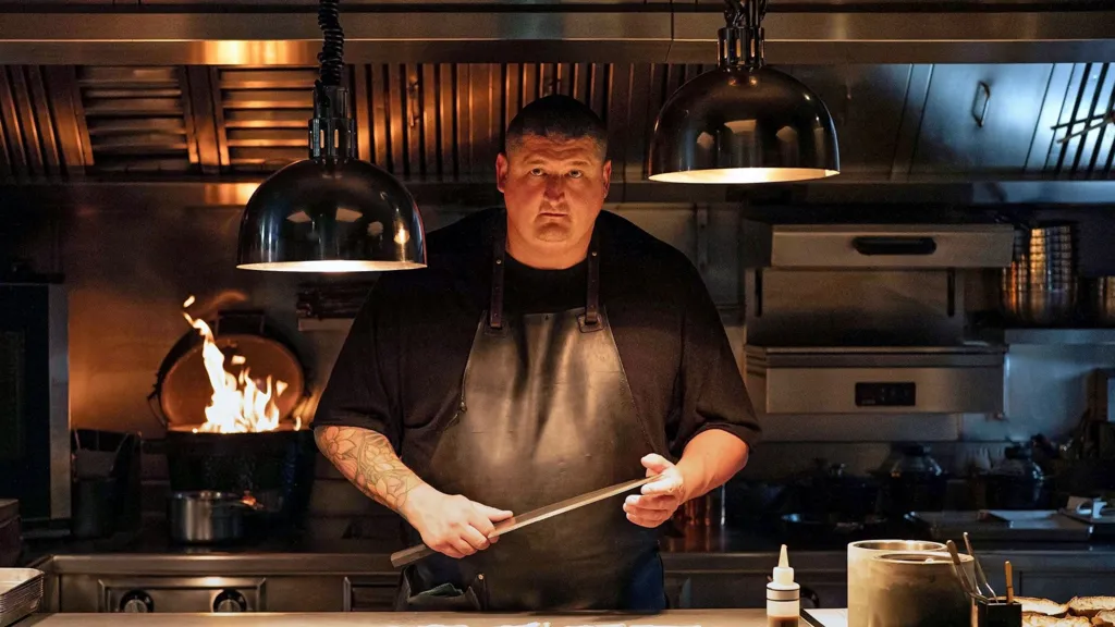 Apple TV Gareth Ward in black chef overalls holding large knife with kitchen appliances and open fire behind him and large overhead light pendants. He has a tattoo down one arm and wears a leather apron and has short dark hair.