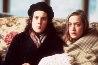 BBC Two people sit closely together on a floral-patterned sofa indoors, wrapped in blankets and warm clothing, facing forward in a softly-lit room.