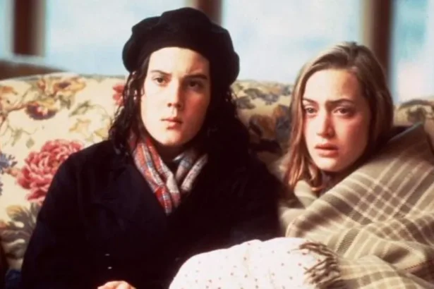 BBC Two people sit closely together on a floral-patterned sofa indoors, wrapped in blankets and warm clothing, facing forward in a softly-lit room.