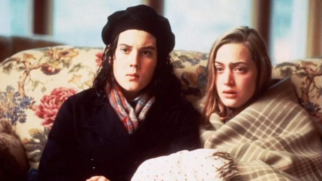 63405b70-d135-11f1-be77-d1864d279cb0-jpg BBC Two people sit closely together on a floral-patterned sofa indoors, wrapped in blankets and warm clothing, facing forward in a softly-lit room.