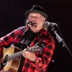 Getty Images Neil young wears a red and black checked shirt and plays a guitar.