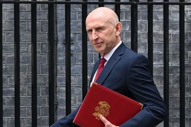 Getty Images Chancellor John Healey walks outside, wearing a navy suit with a red tie. He is holding a red folding and is looking to the side. Behind him is a brick wall and black railings.